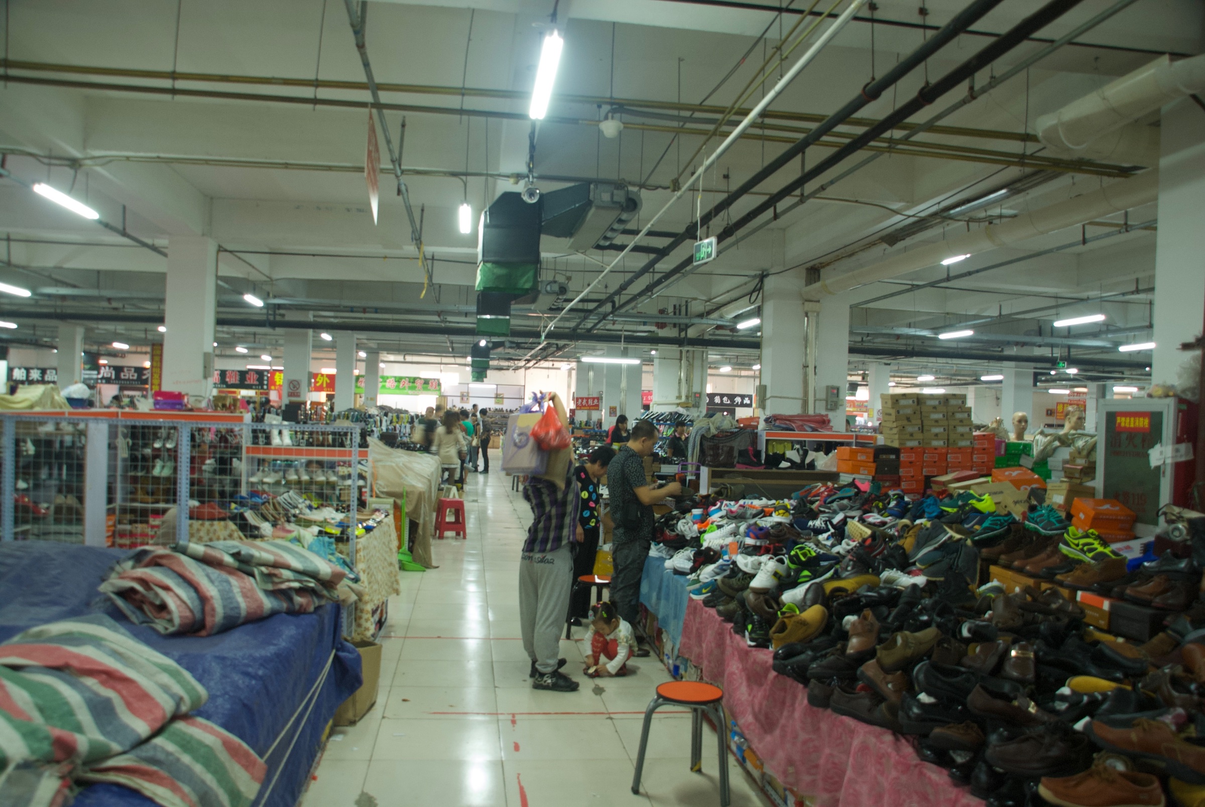 Indoor shoe market with crowded footwear displays, stacked boxes and people along a tiled aisle, documented for Dashilar Flagship Store by Sander Wassink